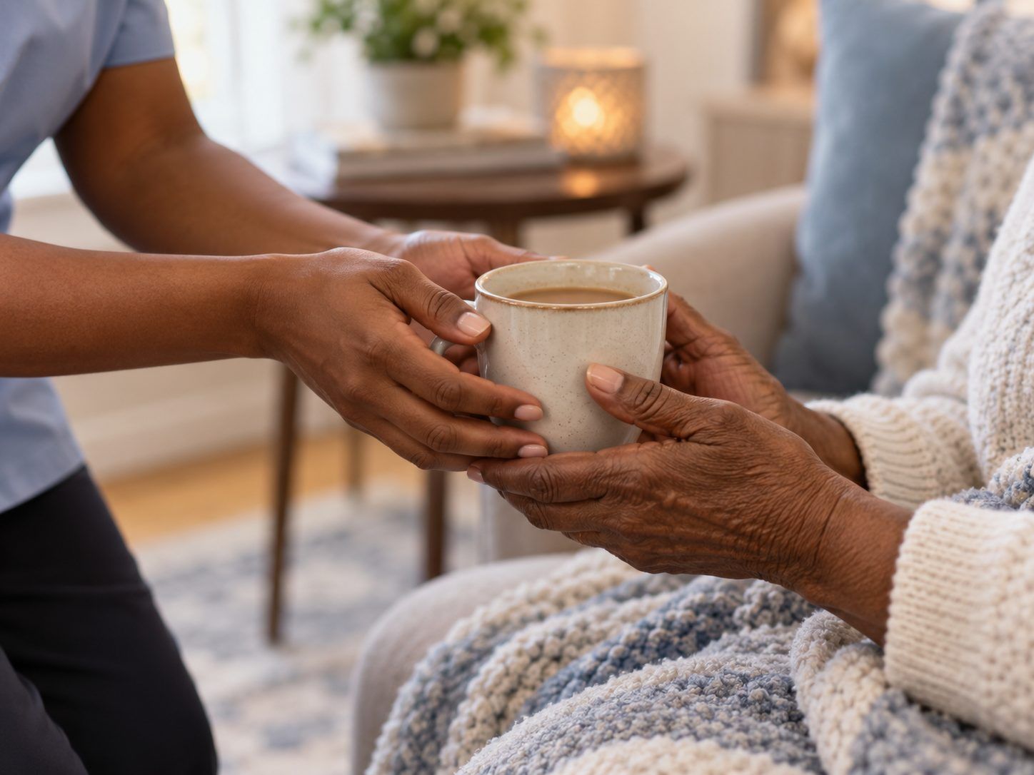 A caregiver's hands passing a mug of coffee to an older person's hands resting on a knitted blanket